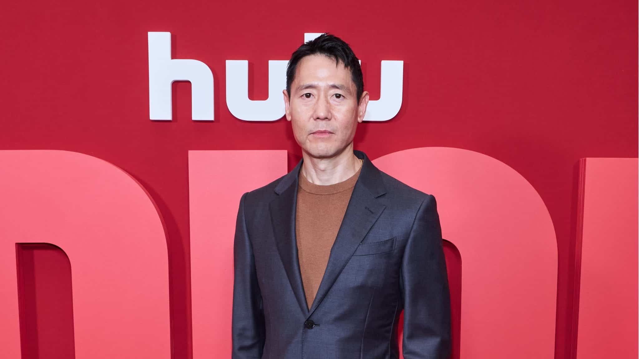 Rob Yang, dressed in a gray suit and brown shirt, stands in front of a red backdrop adorned with the white Hulu logo.