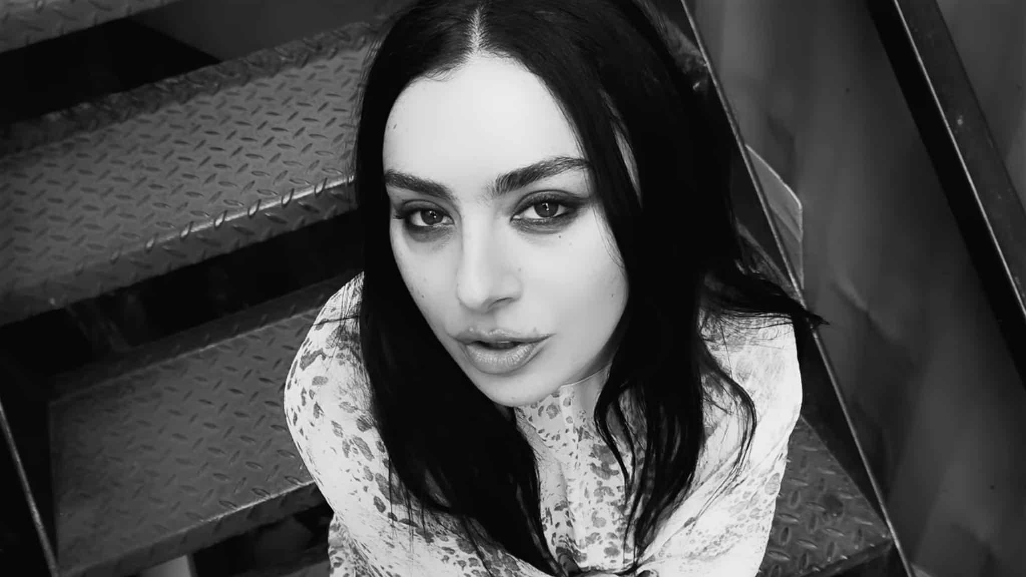 A woman with long dark hair and light eyes looks up at the camera while sitting on metal stairs. She wears a patterned, long-sleeve top, and the photo is in black and white.