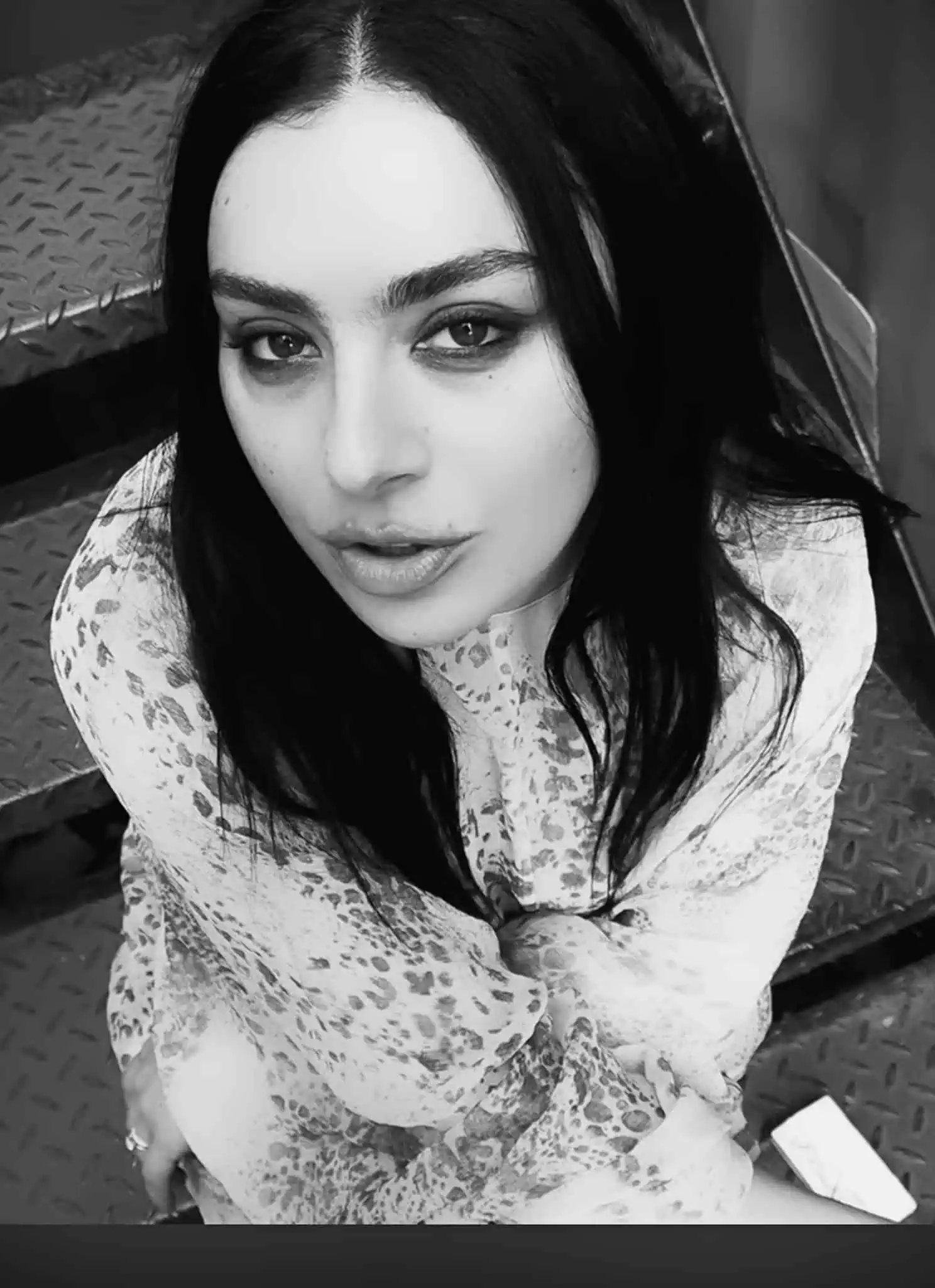 A woman with long dark hair and light eyes looks up at the camera while sitting on metal stairs. She wears a patterned, long-sleeve top, and the photo is in black and white.