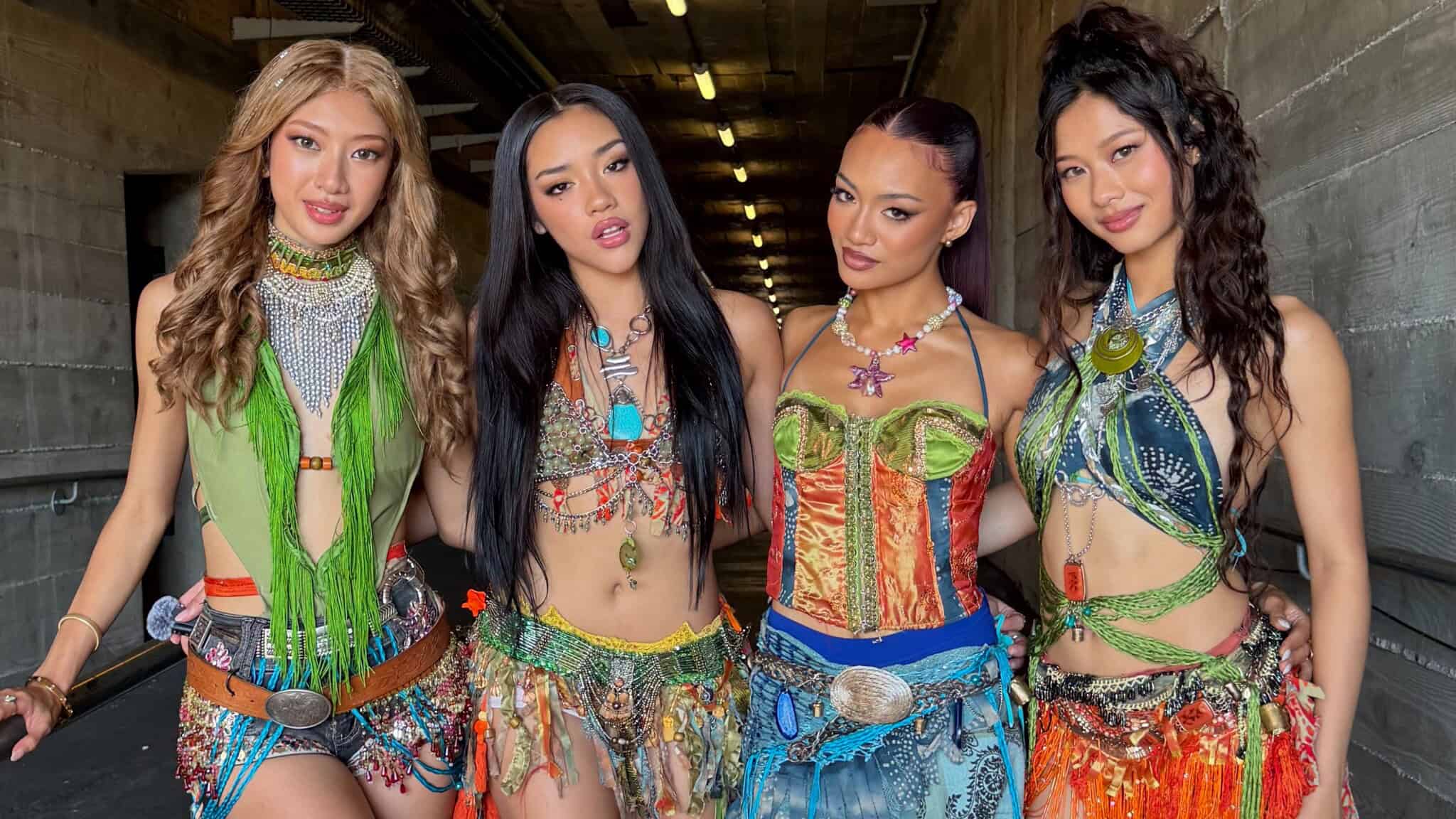 Four women stand side by side in colorful, bohemian outfits with fringe, beads, and jewelry. They pose confidently in a dimly lit hallway with concrete walls and ceiling.