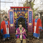Three people in traditional robes pose outdoors in front of a colorful, ornate ceremonial archway decorated with dragons and Chinese characters. The central person is seated, while the other two stand on either side.