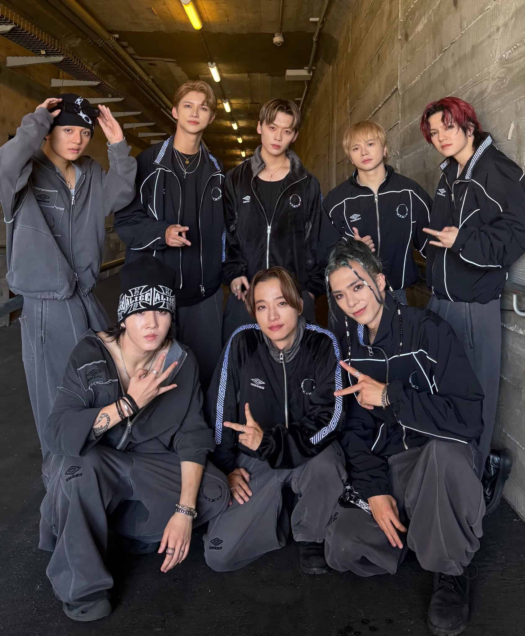 Eight people pose together in a concrete hallway, wearing coordinated black and gray outfits. Some are standing, others crouching, and several are making peace signs or playful gestures for the camera.