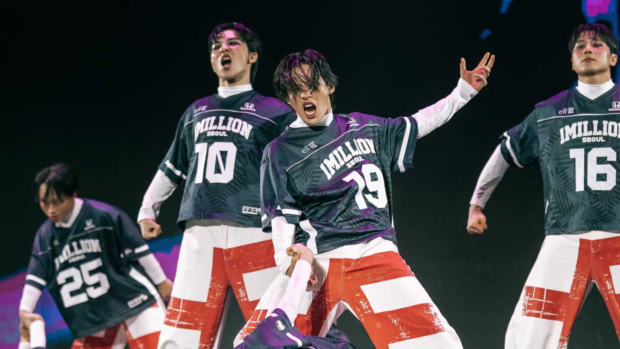Five dancers in matching black jerseys and red-and-white pants passionately perform on stage, striking dynamic poses with intense facial expressions.