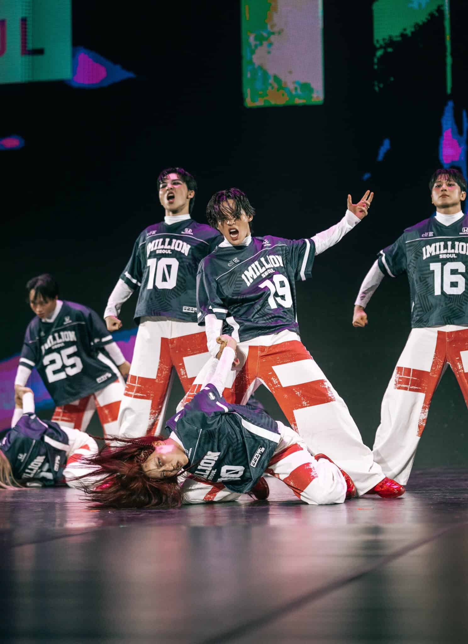 Five dancers in matching black jerseys and red-and-white pants passionately perform on stage, striking dynamic poses with intense facial expressions.