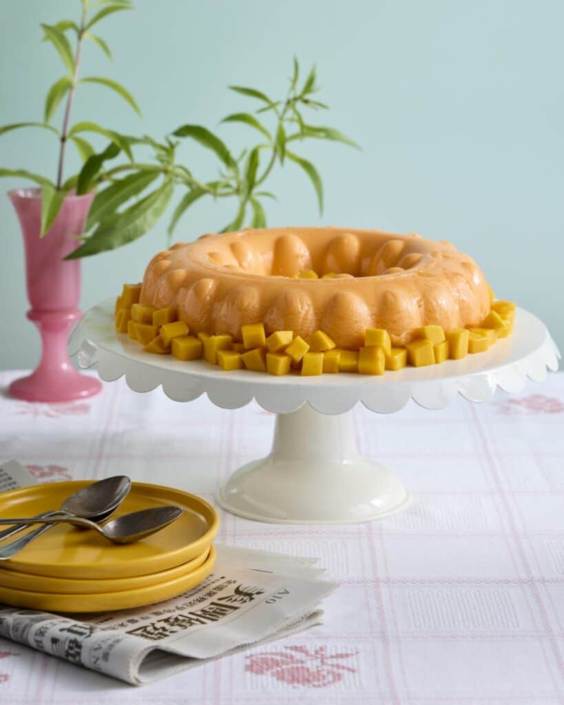 A mango gelatin mold on a white cake stand, surrounded by mango cubes. Nearby are stacked yellow plates with spoons, a newspaper, and a leafy plant in a pink vase on a pink-and-white checkered tablecloth.