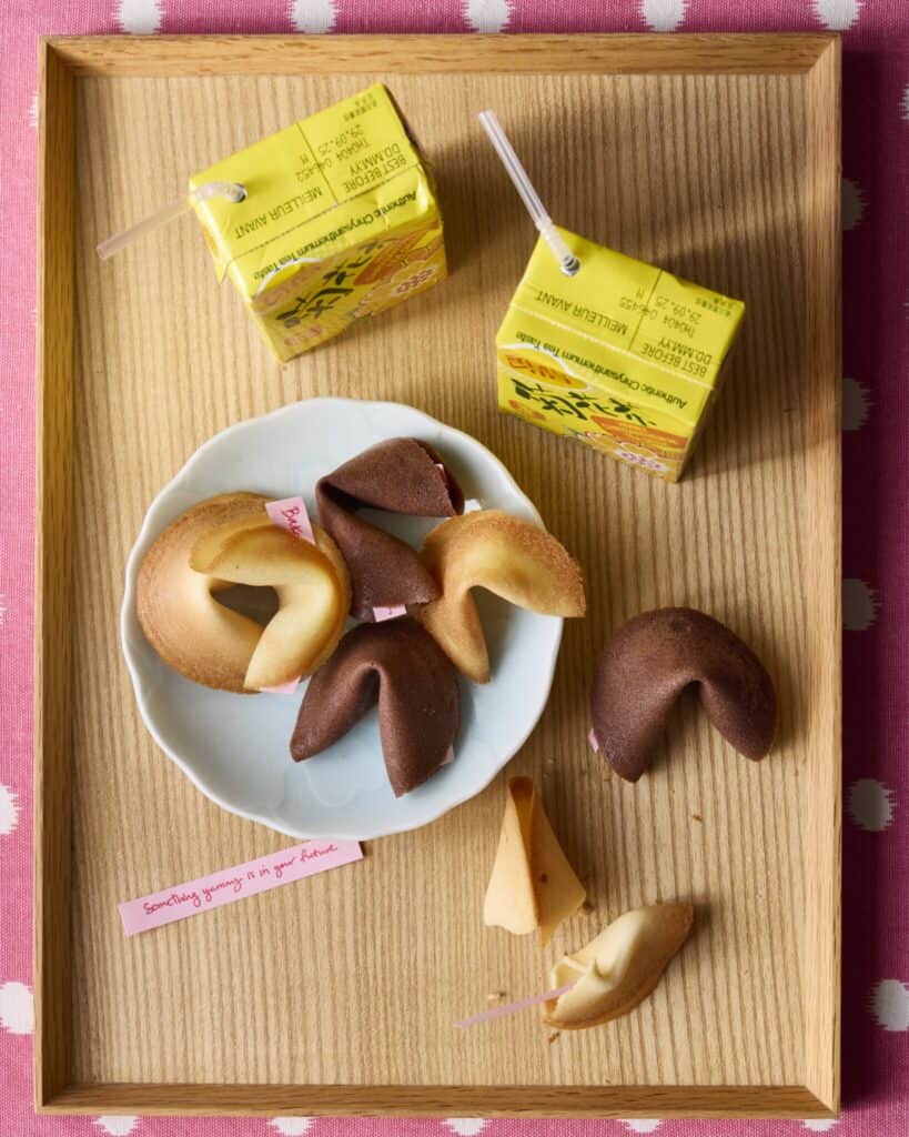 A wooden tray with fortune cookies, some chocolate-flavored, on a plate and around it, plus two small yellow juice boxes with straws, all set on a pink cloth with white dots. Some fortunes are visible.