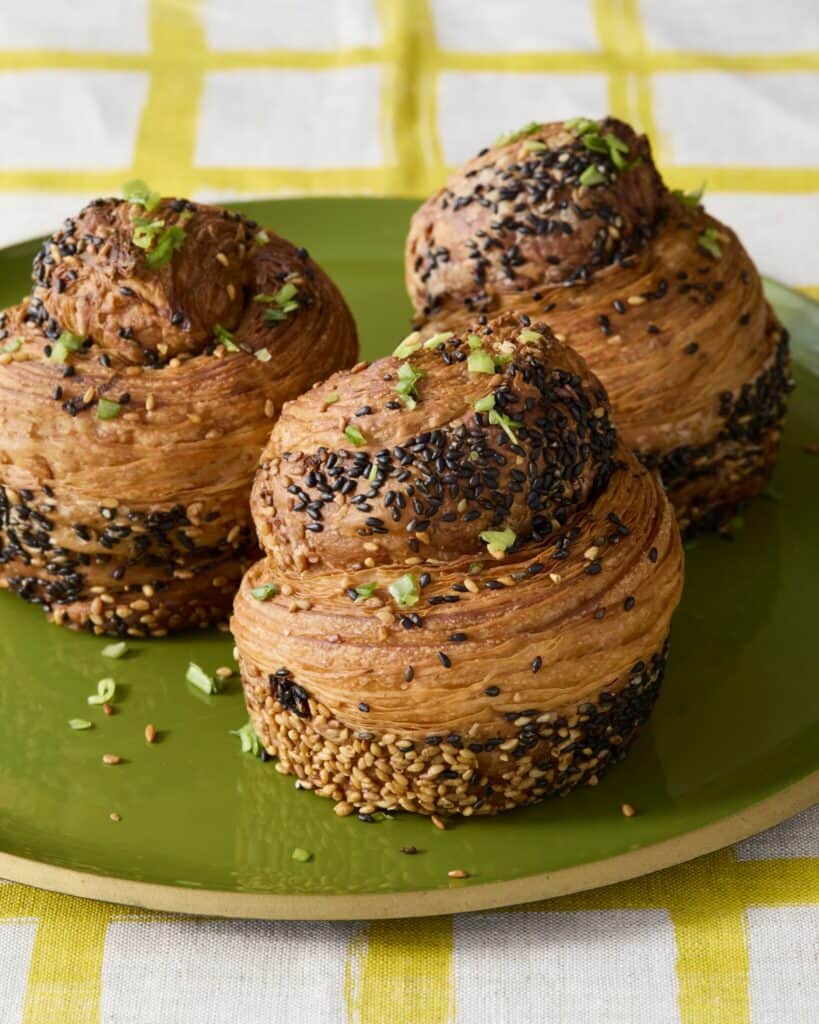 Three flaky, golden pastry rolls topped with black sesame seeds and chopped herbs are arranged on a green plate, set on a yellow and white checkered tablecloth.