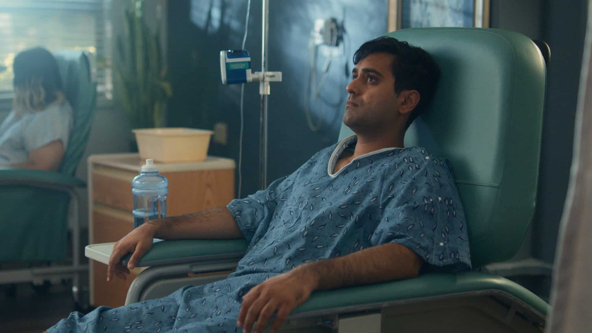 A man in a hospital gown sits in a reclining medical chair, receiving IV treatment. He looks thoughtful. A water bottle is on the table next to him, and another patient is visible in the background.
