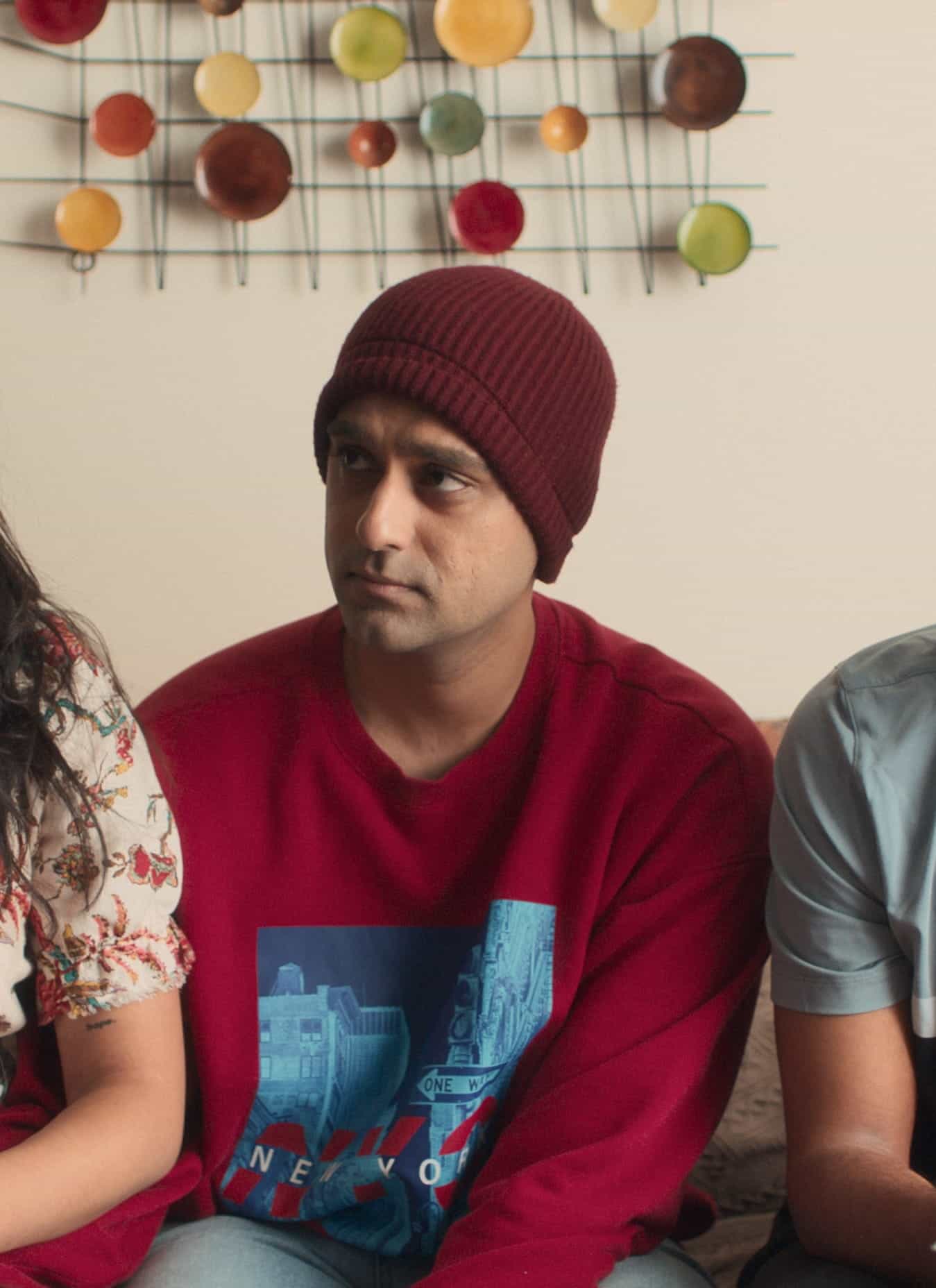 Three people sit on a couch looking thoughtful or concerned. A woman in a floral dress is on the left, a man in a red beanie and sweater is in the middle, and another man holds a plate on the right. Colorful wall art hangs behind them.
