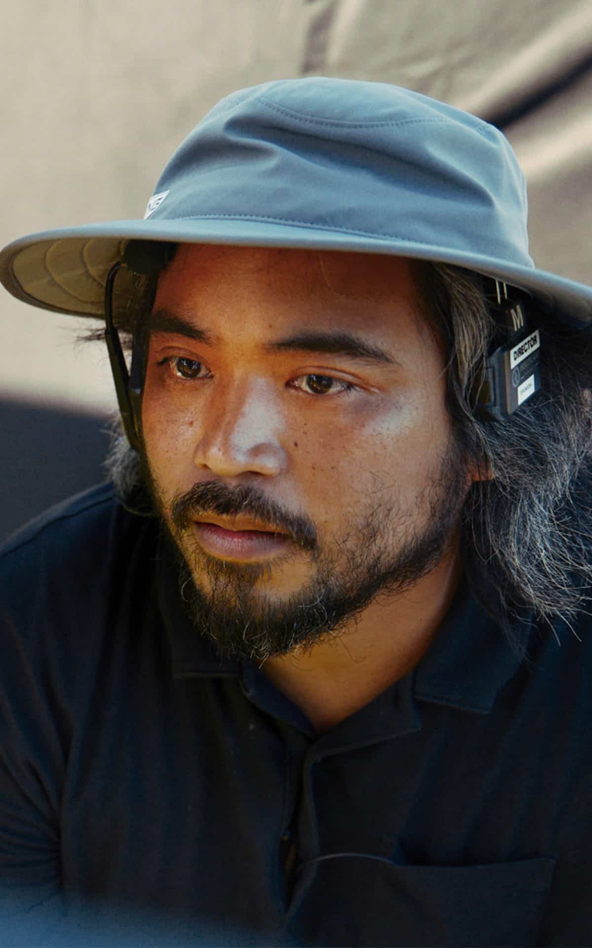 A man with a beard and mustache wears a gray wide-brimmed hat and black shirt. Reminiscent of Filipiñana style, he has headphones on and looks focused, with a neutral background behind him—capturing the contemplative essence often seen in Rafael Manuel’s portraits.