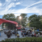 People sit on chairs in an outdoor plaza, watching a large screen under a modern red canopy, as the sun sets behind trees and clouds in the background.