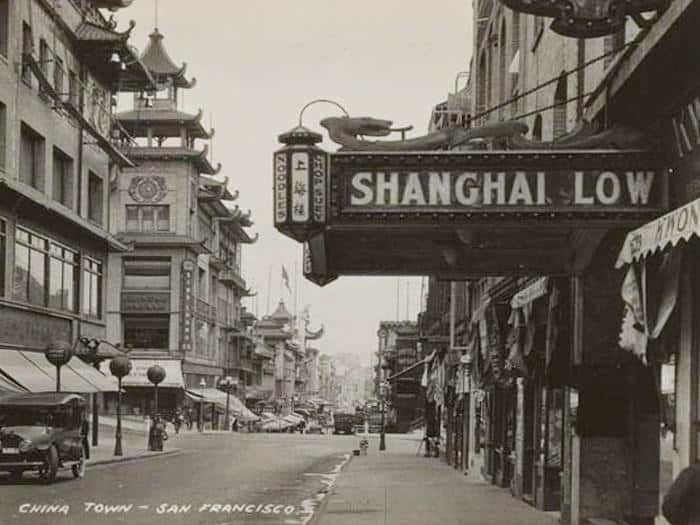 Black-and-white photo of San Franciscos Chinatown, featuring a street scene with cars, buildings with Chinese architectural details, and a prominent Shanghai Low restaurant sign.