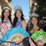 Three women in crown tiaras and sashes stand outdoors in a festive street. Two women wear floral dresses and hold blue and green fans. Lanterns are visible in the background.