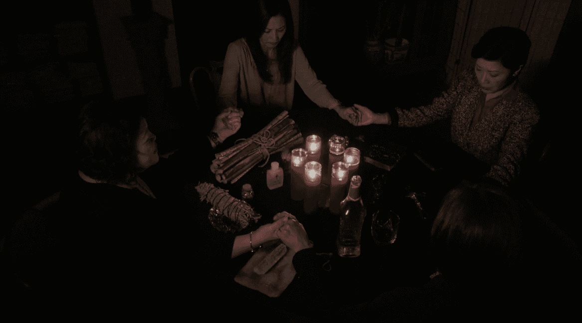Four women sit around a dimly lit table holding hands, surrounded by red candles, a bottle, and ritual objects, creating a solemn, ritualistic atmosphere.