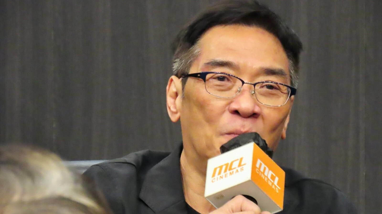 A man with glasses and short dark hair speaks into an orange microphone labeled MCL CINEMAS against a dark background.