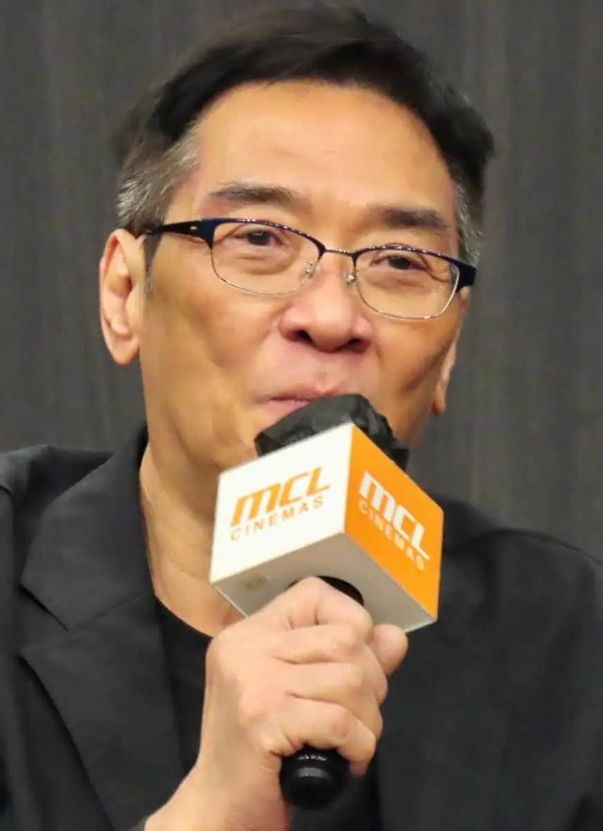 A man with glasses and short dark hair speaks into an orange microphone labeled MCL CINEMAS against a dark background.