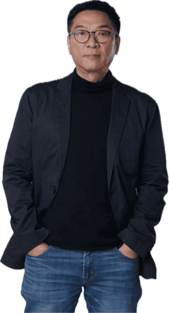 A man wearing glasses, a black blazer, a black turtleneck, and blue jeans stands with his hands in his pockets against a white background.