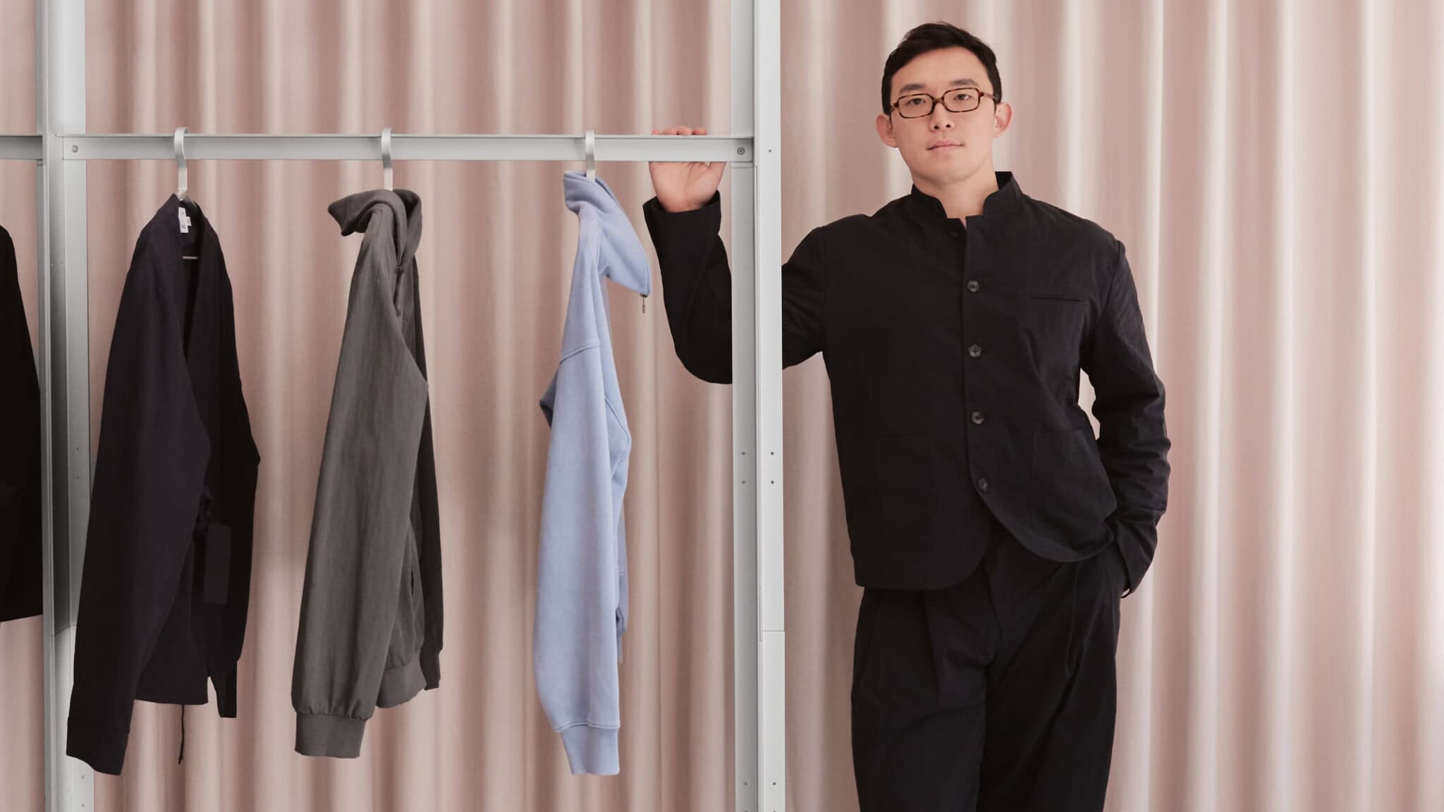 A person in black clothing stands by a clothing rack with three hanging garments: a black jacket, a gray sweatshirt, and a light blue hoodie. Pale pink curtains are in the background.