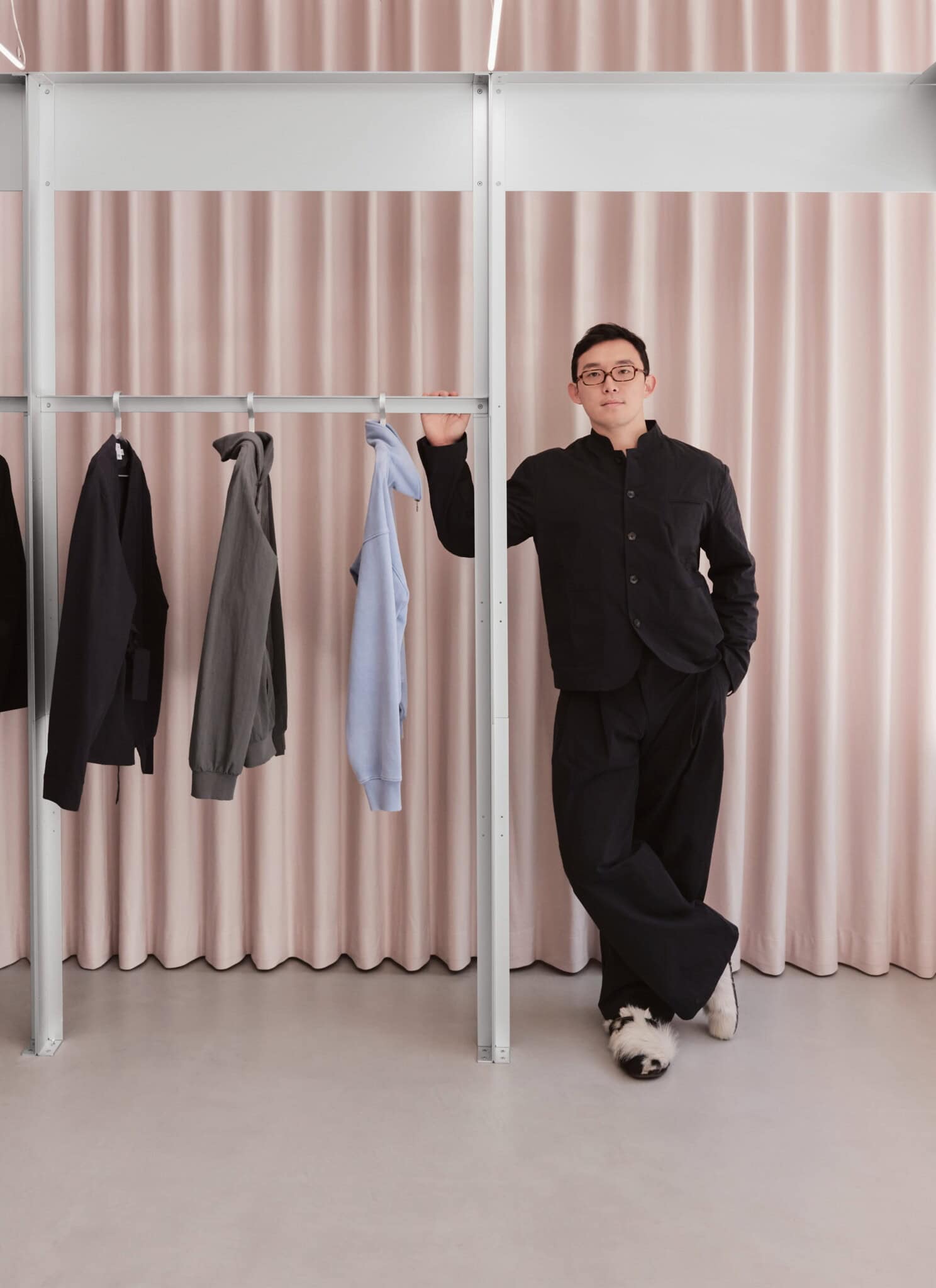 A person in black clothing stands by a clothing rack with three hanging garments: a black jacket, a gray sweatshirt, and a light blue hoodie. Pale pink curtains are in the background.