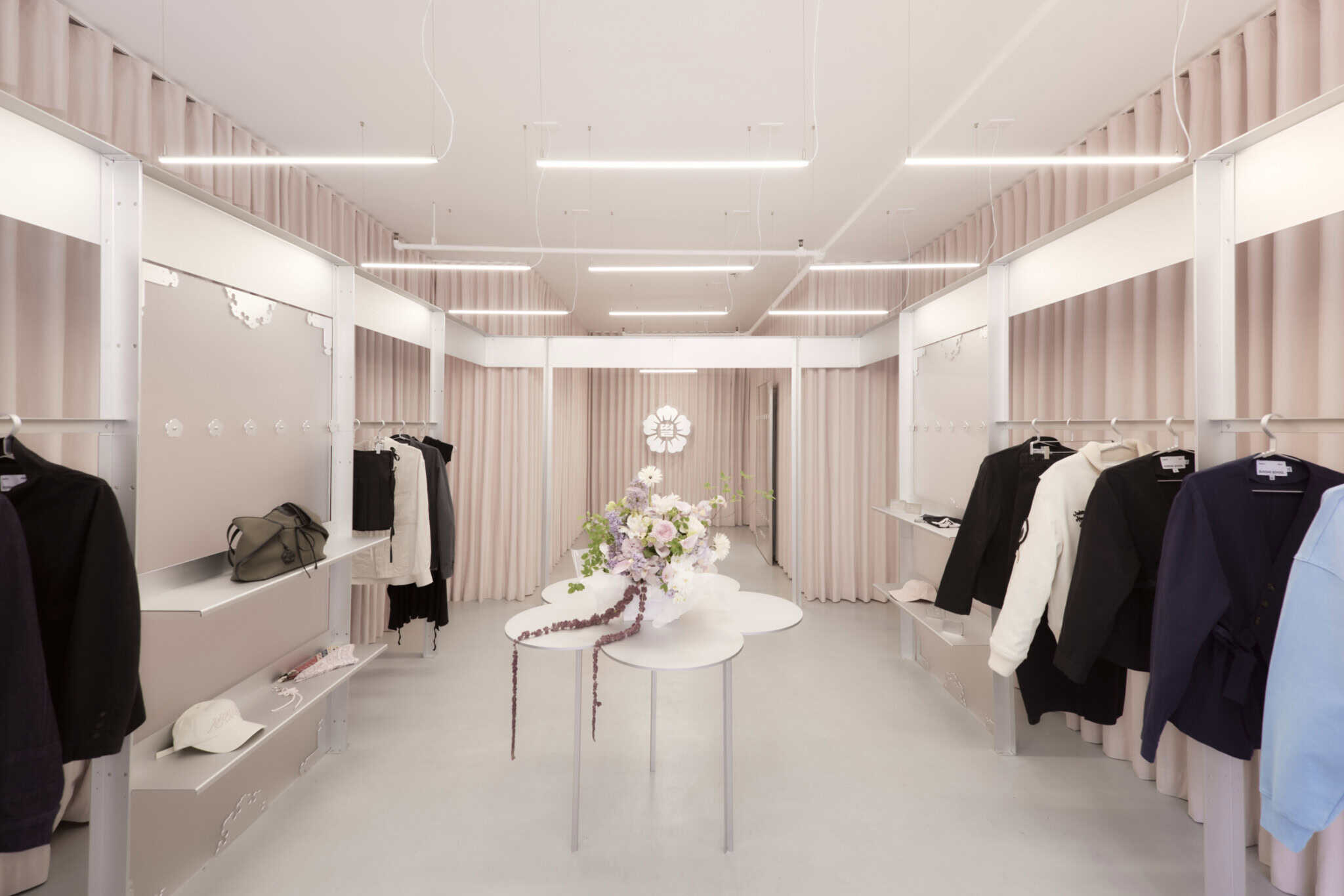Minimalist clothing boutique with soft pink walls and shelves, round white tables with flower arrangements in the center, and various garments hanging neatly on both sides. Bright ceiling lights illuminate the space.
