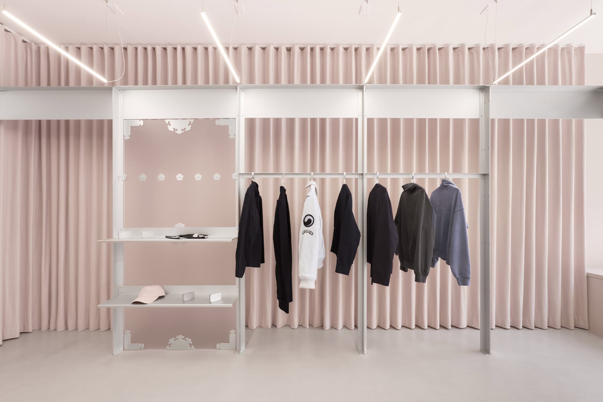 Minimalist clothing store with pink curtains, white shelves, and a silver rack displaying black, gray, and one white hoodie. Soft lighting and clean lines create a modern, elegant atmosphere.