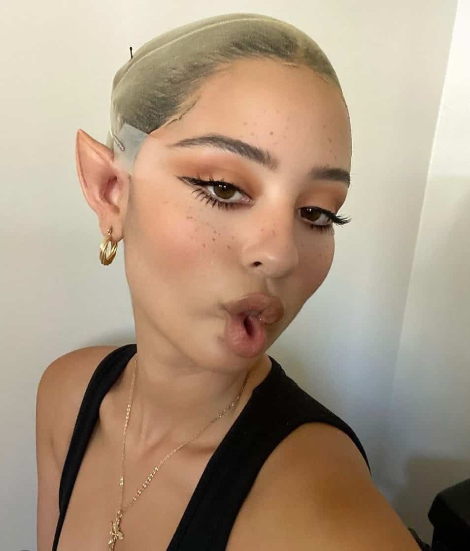 A person wearing a wig cap and pointed elf ears makes a playful fish face, with winged eyeliner, freckles, gold hoop earrings, and a necklace, posing for a selfie against a plain background.