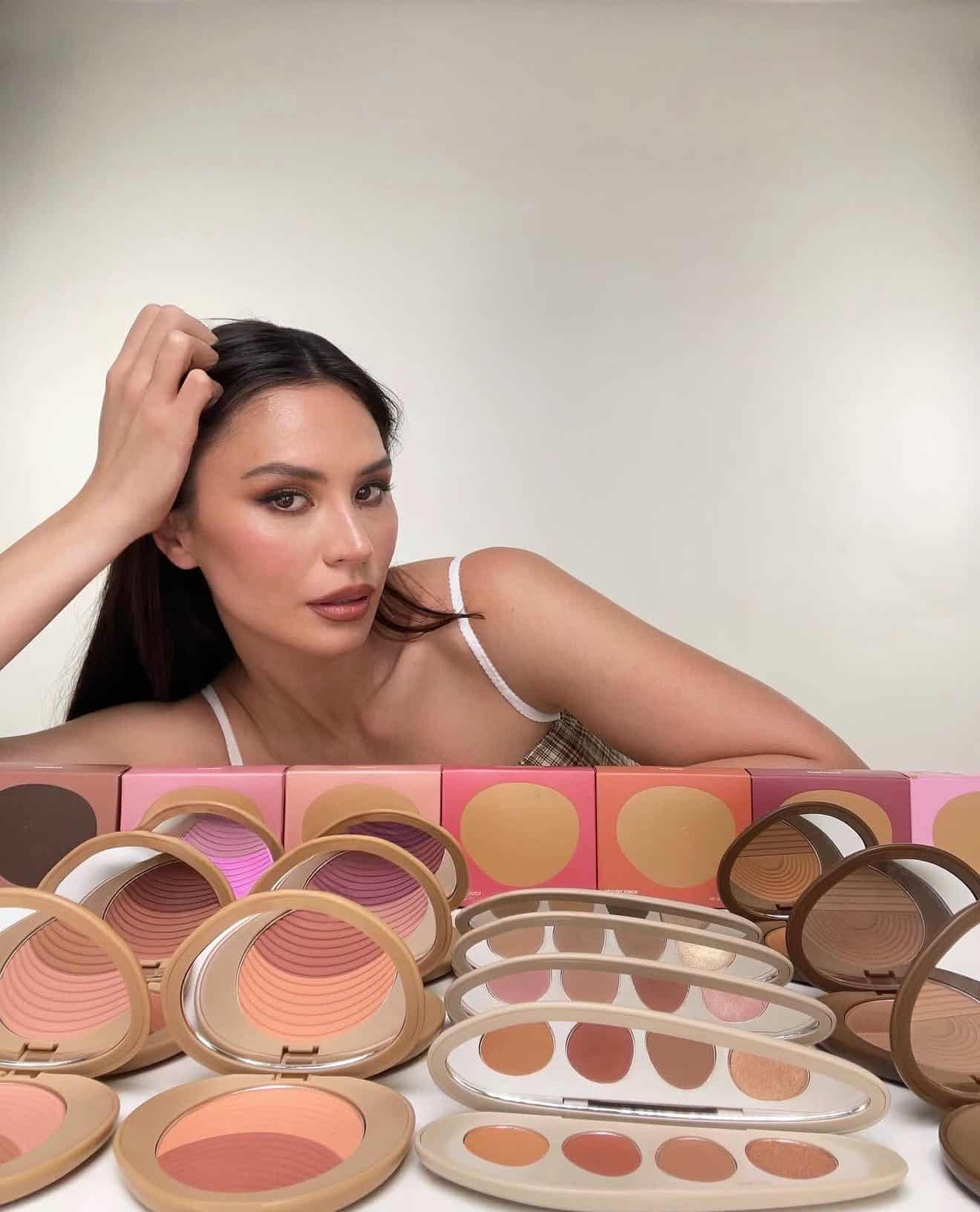 A woman with long dark hair poses behind various open makeup compacts displaying powders and eyeshadows in different shades, set against a plain background.