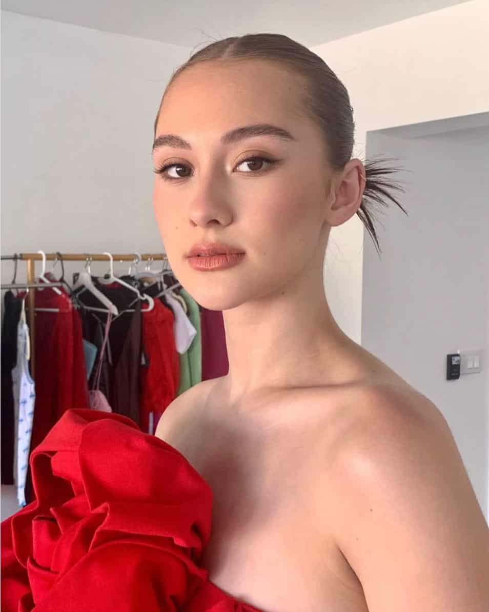 A person with neatly tied-back hair and natural makeup wears a red off-shoulder outfit. Clothing racks with various garments are in the background, and the setting appears to be a dressing room.