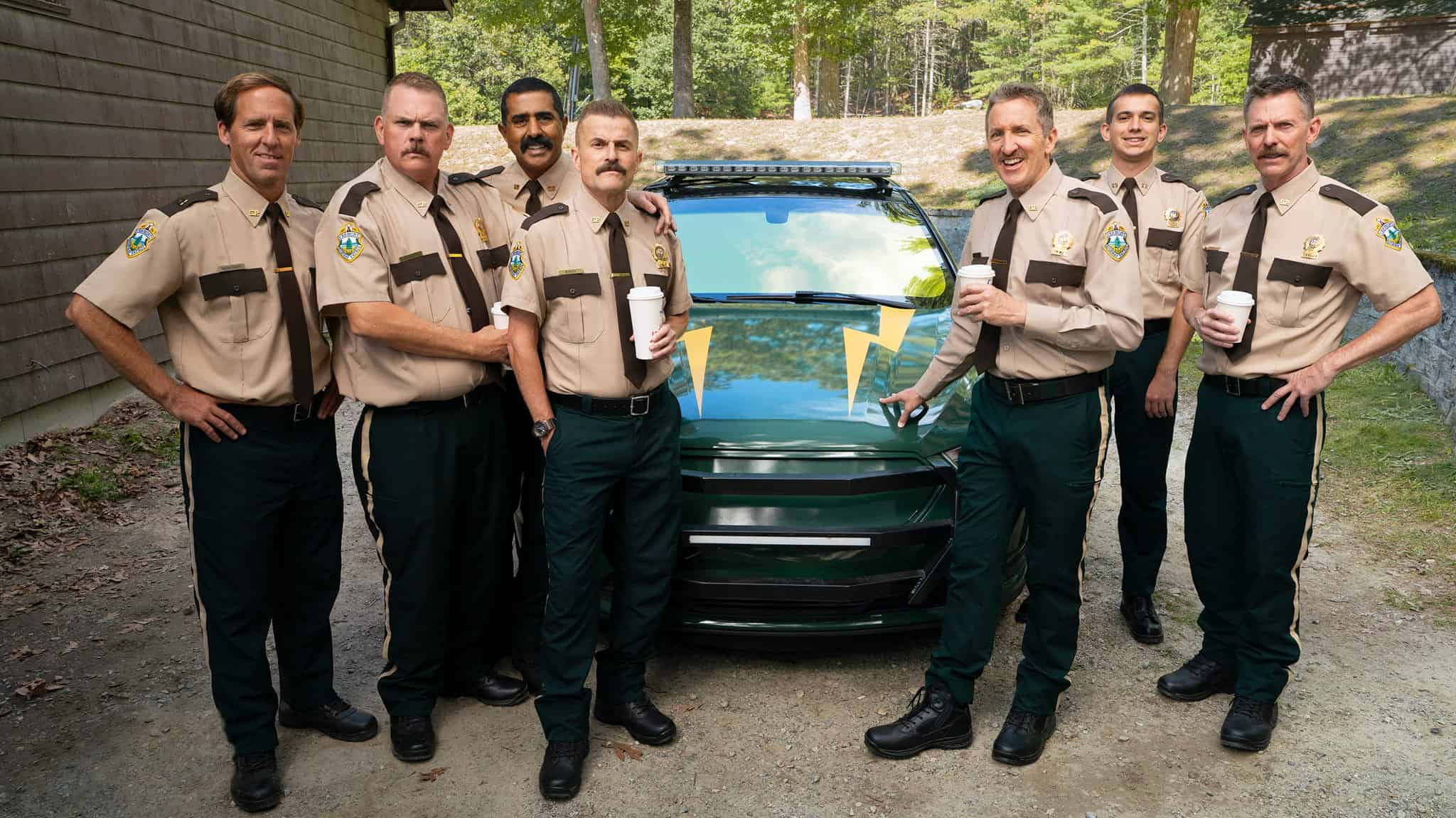 Seven uniformed officers stand smiling in front of a green police car in a wooded area, holding coffee cups. They are wearing tan shirts, dark pants, and badges, appearing relaxed and cheerful together.