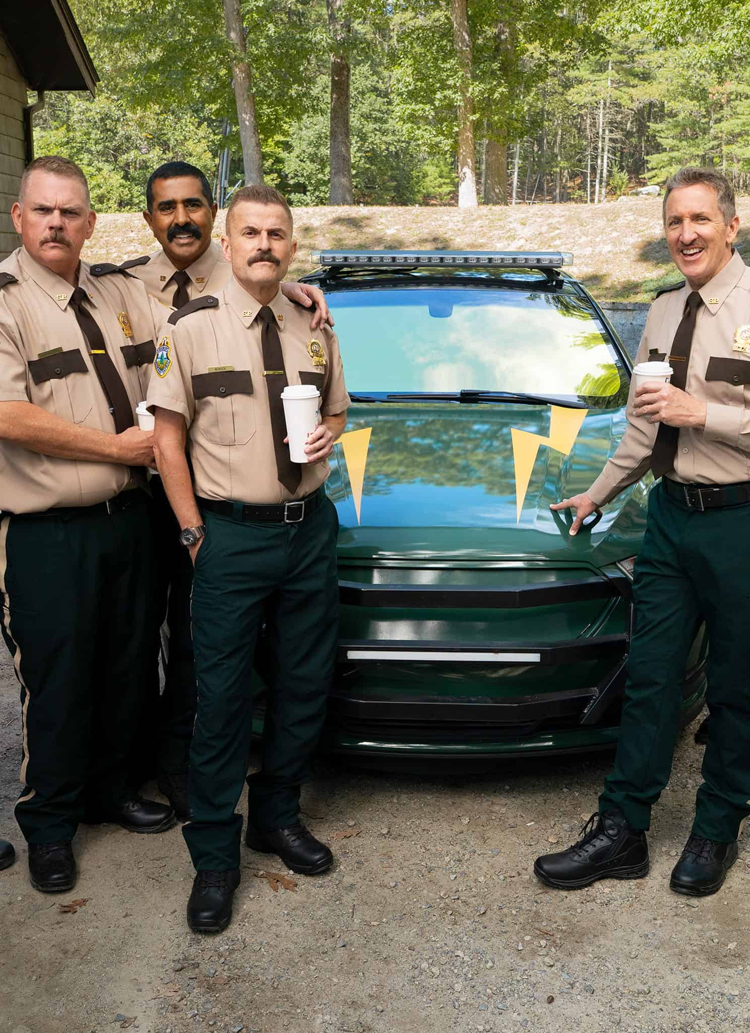 Seven uniformed officers stand smiling in front of a green police car in a wooded area, holding coffee cups. They are wearing tan shirts, dark pants, and badges, appearing relaxed and cheerful together.