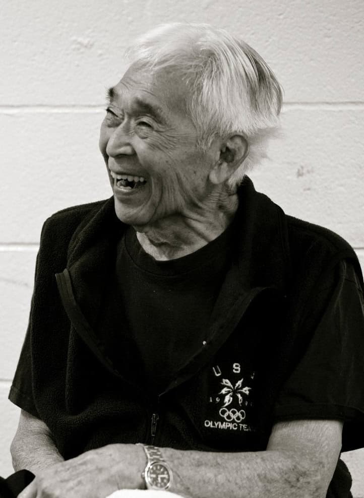 An elderly man with white hair, identified as Tommy Kono, smiles and laughs while sitting. He wears a dark jacket and a T-shirt with USA Olympic Team printed on it. The background is a plain, light-colored wall.