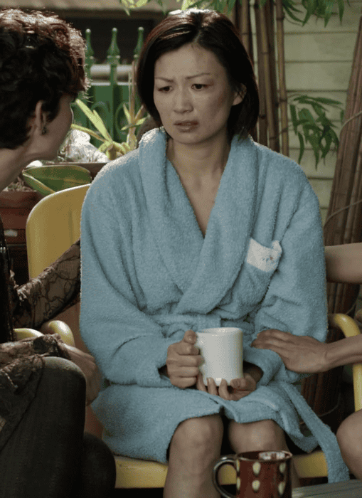 Three women sit together outdoors. The woman in the center, wearing a blue bathrobe and holding a mug, looks upset. The women on both sides gently comfort her by placing their hands on her arms, creating a quiet moment of support in the presence of The Unbidden.