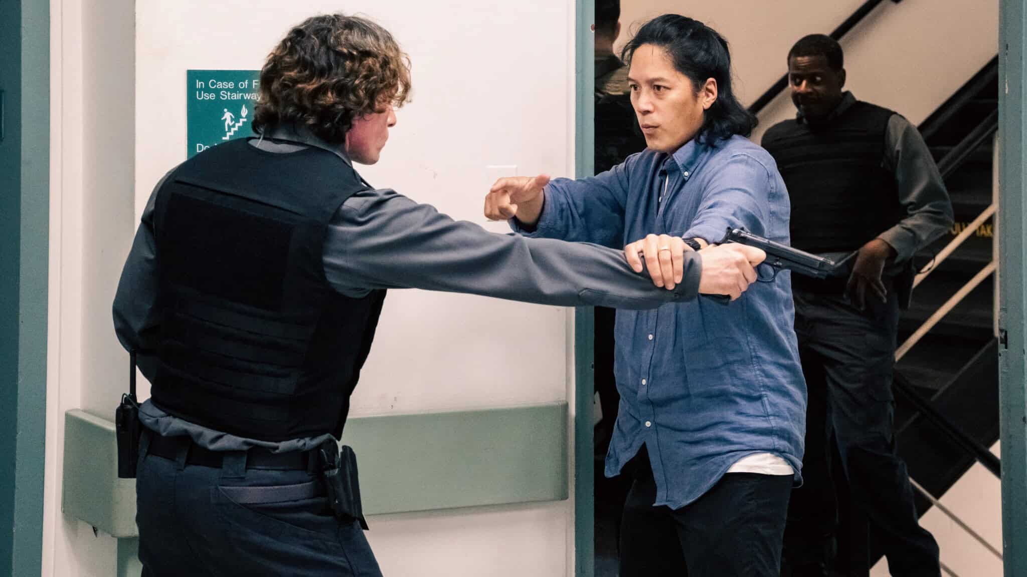 A person in a blue shirt confronts a uniformed officer with a gun in a tense hallway scene, while others in tactical vests watch from behind near a stairwell.