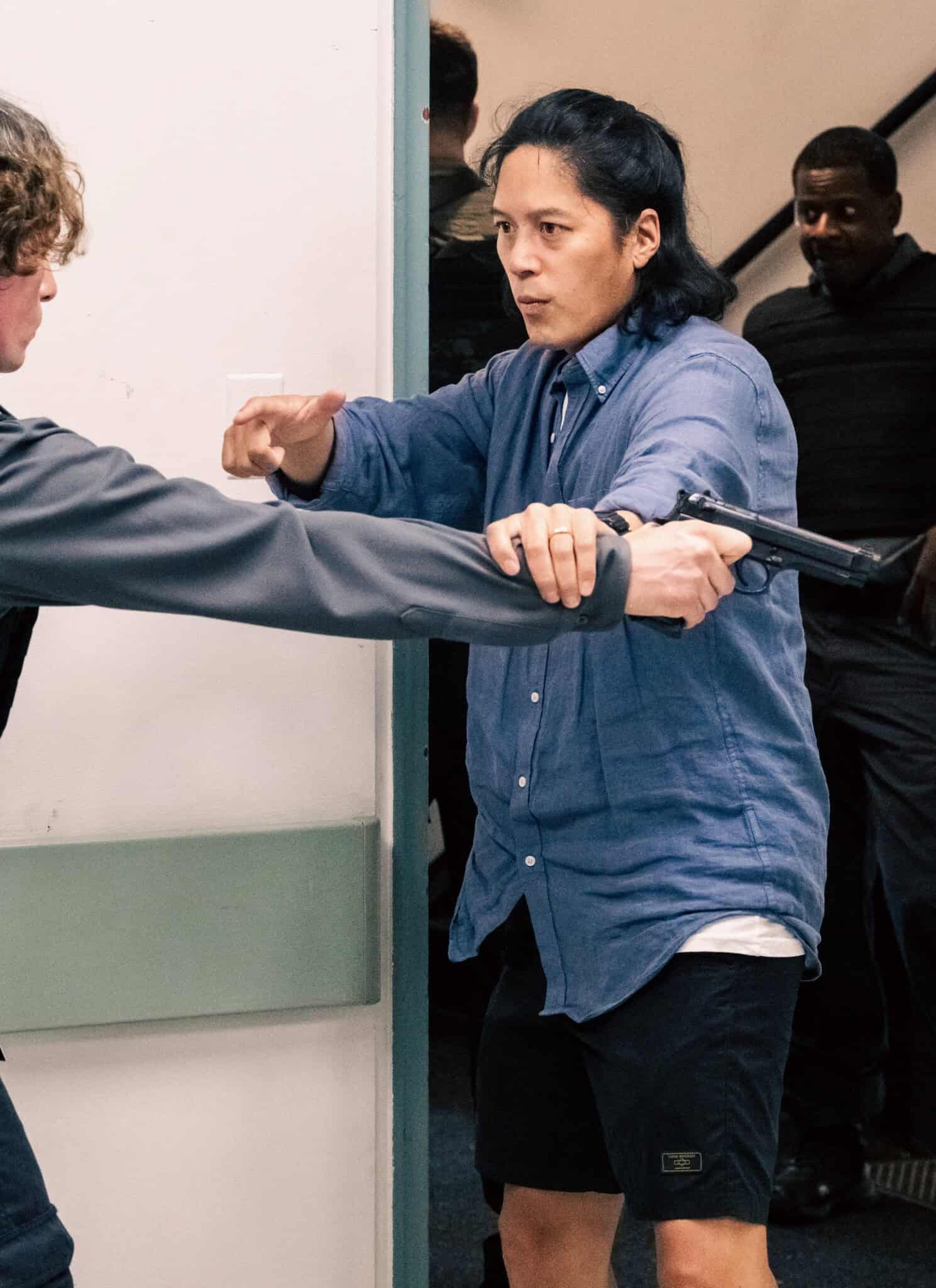 A person in a blue shirt confronts a uniformed officer with a gun in a tense hallway scene, while others in tactical vests watch from behind near a stairwell.