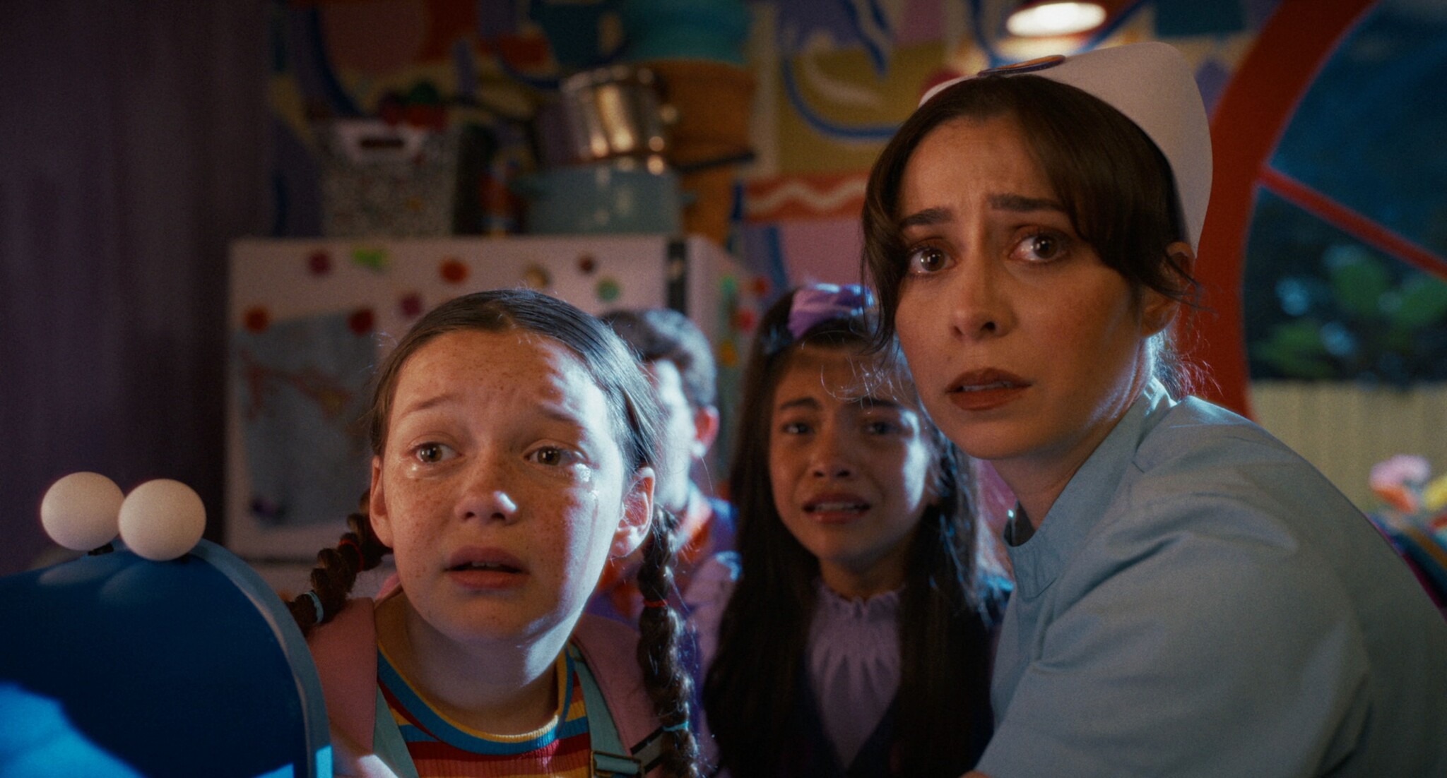 Three girls look frightened while a woman in a nurse outfit, also looking scared, sits beside them. The colorful room behind them has playful decorations and a fridge covered in artwork.