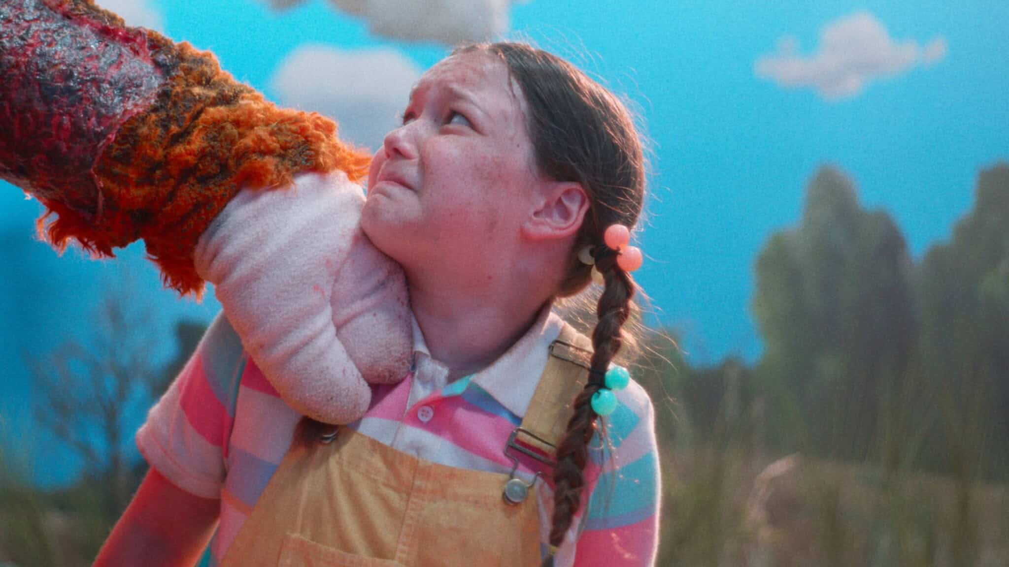 A young girl in striped shirt and yellow overalls looks upset as a large, furry puppet hand grips her collar. The background is bright and outdoorsy with clouds and trees.