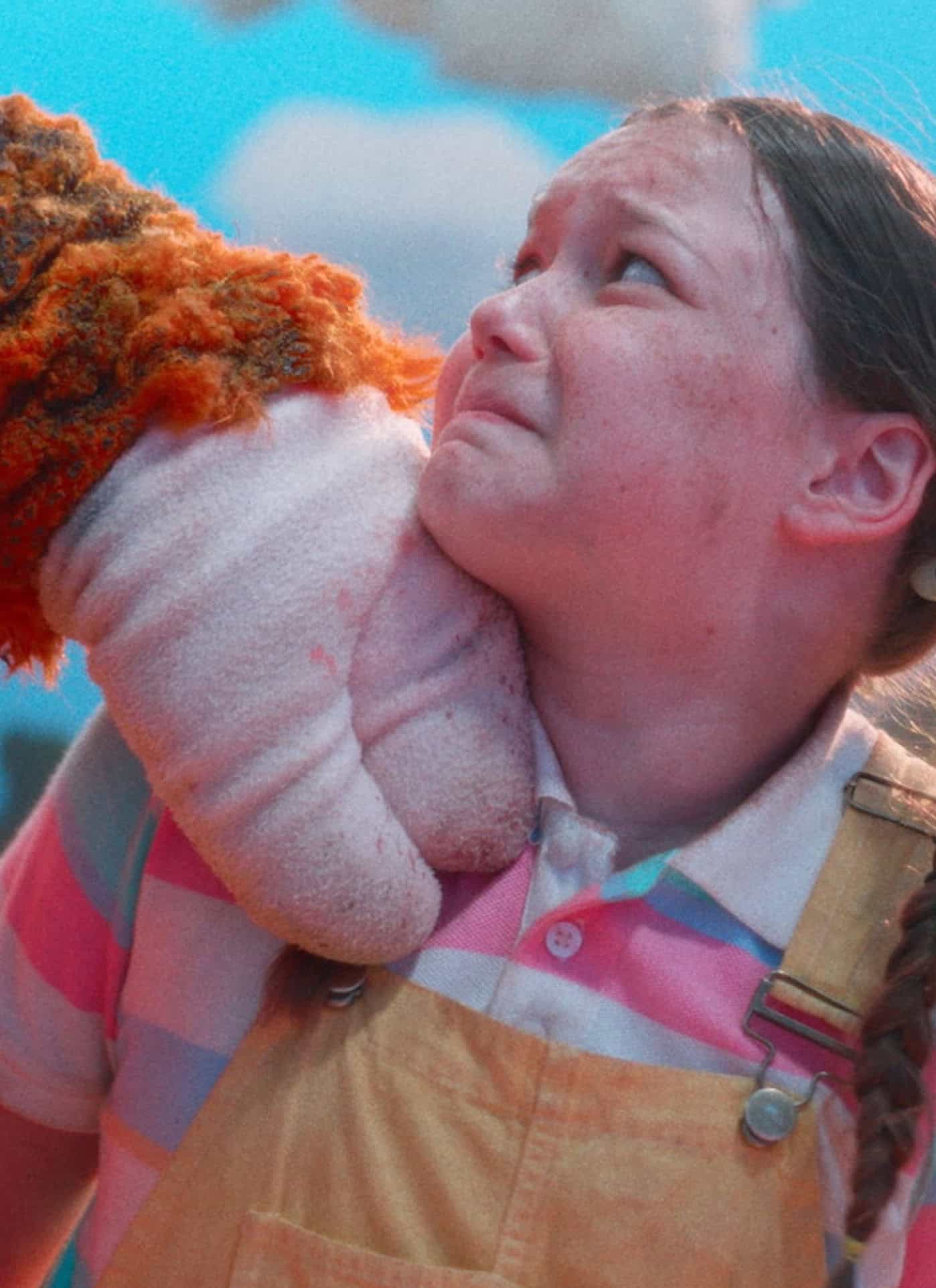 A young girl in striped shirt and yellow overalls looks upset as a large, furry puppet hand grips her collar. The background is bright and outdoorsy with clouds and trees.