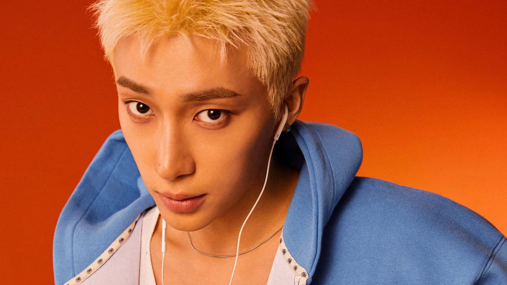 A person with short blond hair wearing a blue hoodie and white earphones looks directly at the camera against an orange background.