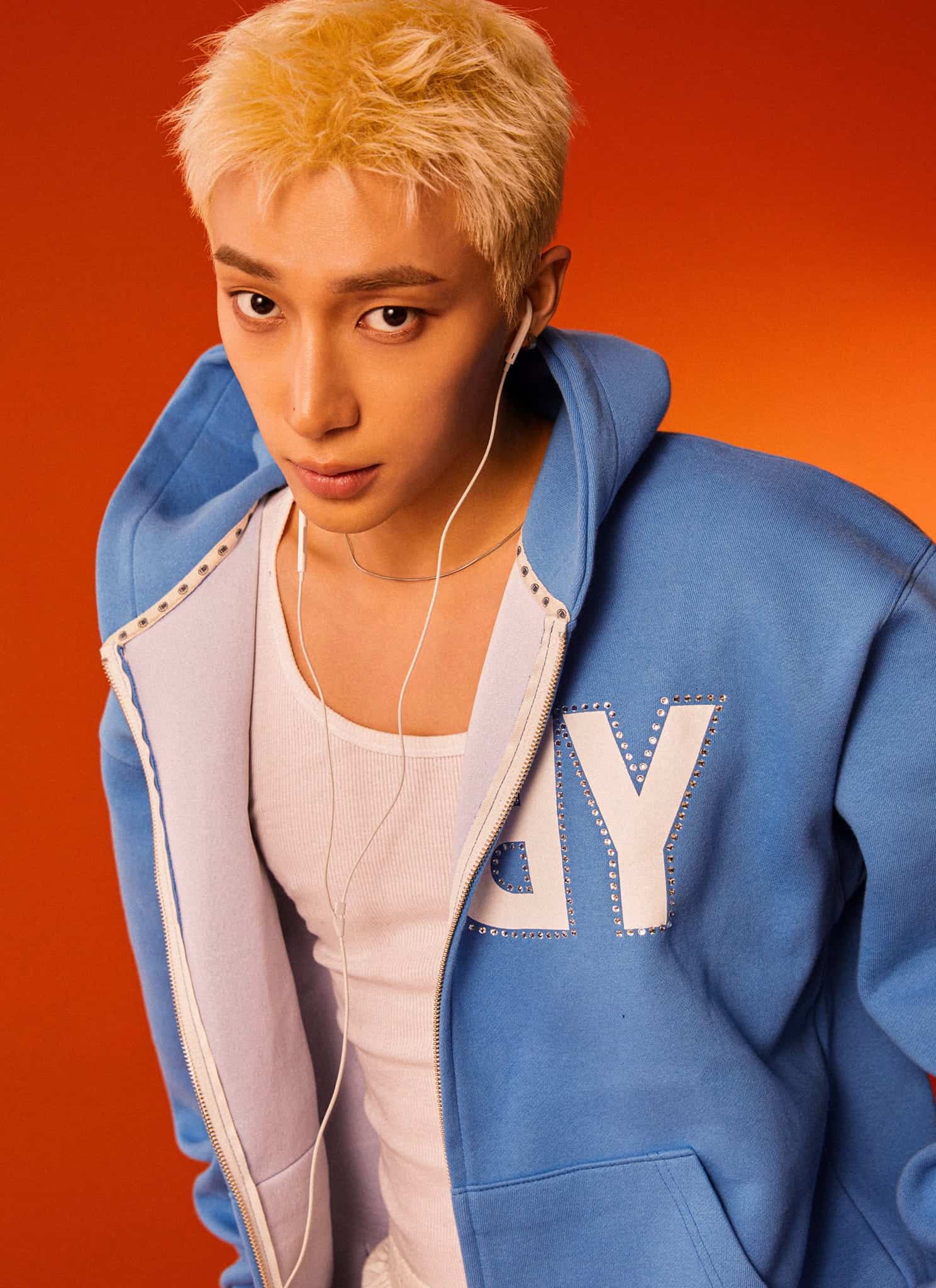 A person with short blond hair wearing a blue hoodie and white earphones looks directly at the camera against an orange background.