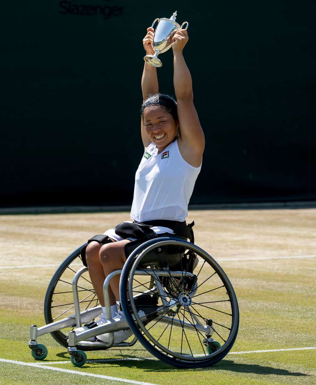 A female wheelchair tennis player smiles and lifts a trophy above her head while sitting on a grass tennis court at Wimbledon.