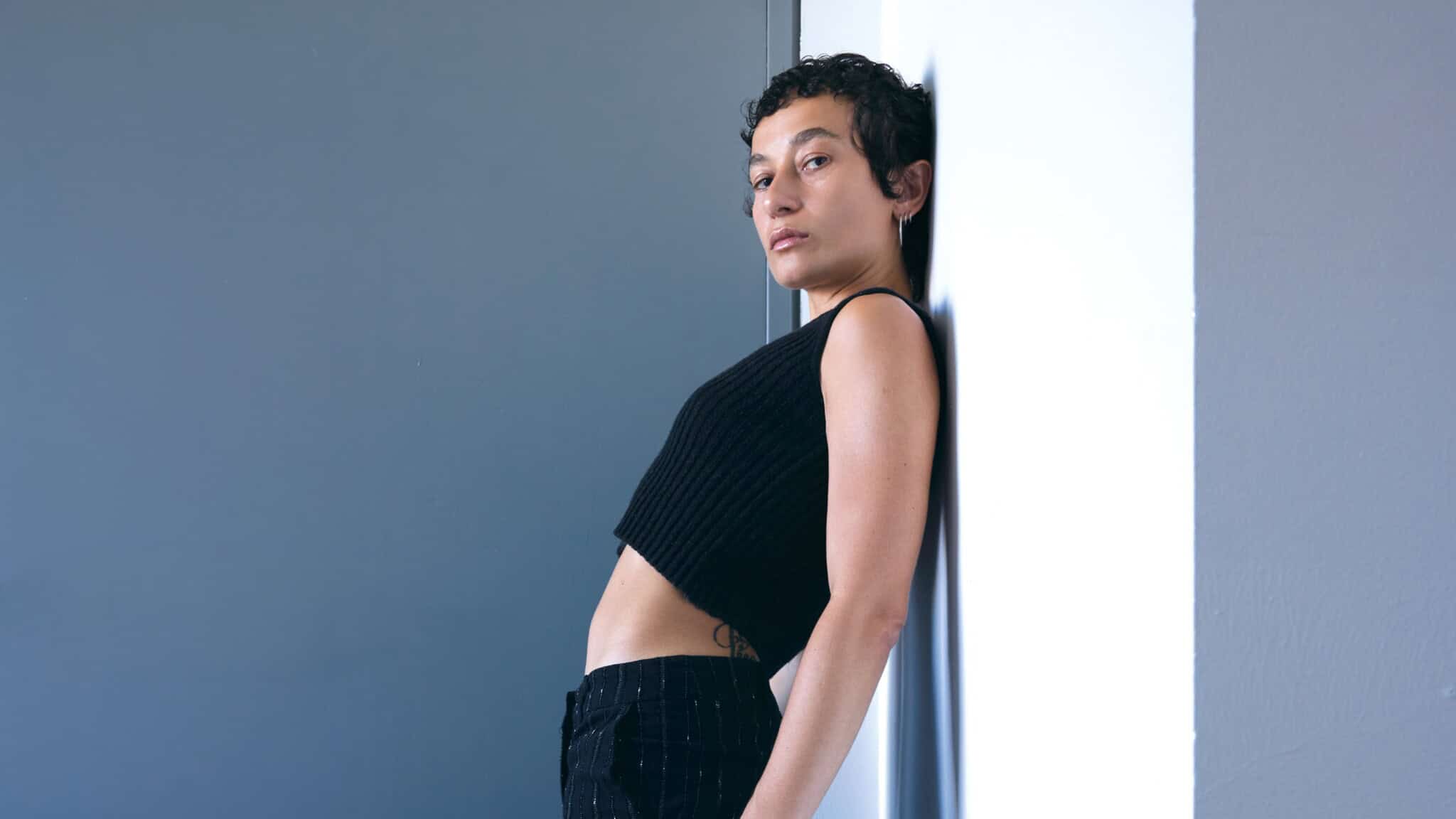 Person with short curly hair, wearing a black sleeveless top and black pants, leans back against a white wall, looking confidently at the camera. Channeling the unmistakable style of Nene H, the background features a modern gray and white color scheme.