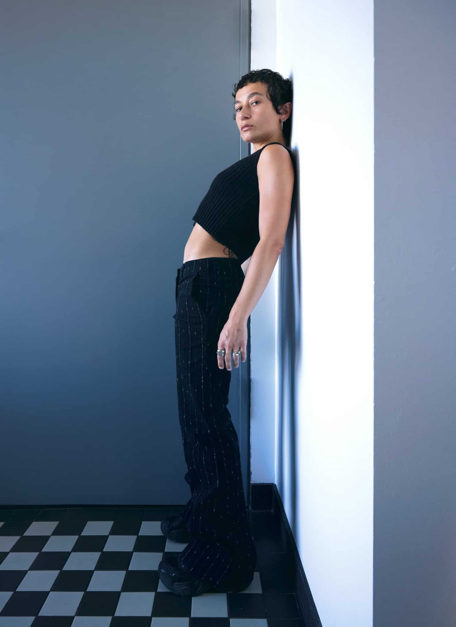 Person with short curly hair, wearing a black sleeveless top and black pants, leans back against a white wall, looking confidently at the camera. Channeling the unmistakable style of Nene H, the background features a modern gray and white color scheme.