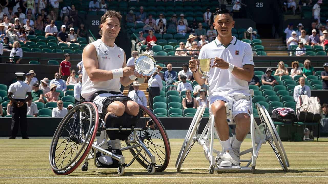 Two male wheelchair tennis players pose on a grass court at Wimbledon, holding their trophies. They are smiling with an audience seated in the background on a sunny day.