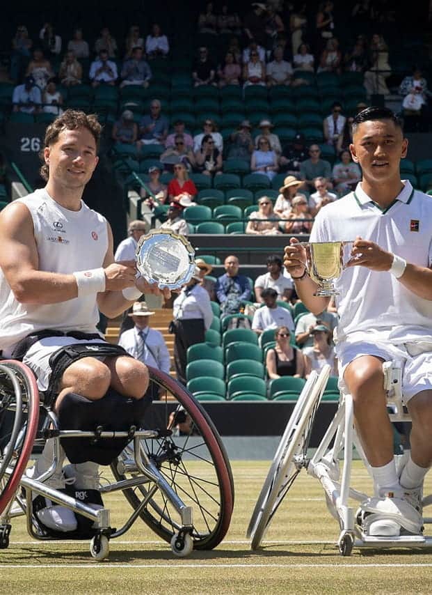 Two male wheelchair tennis players pose on a grass court at Wimbledon, holding their trophies. They are smiling with an audience seated in the background on a sunny day.