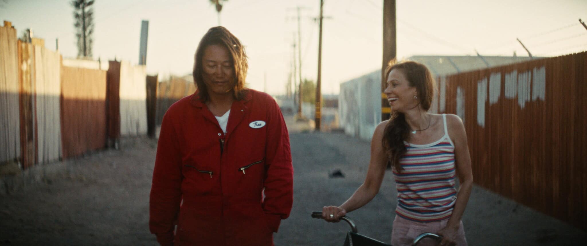 Two people walk together down a sunlit alley; one wears a red jumpsuit, the other smiles while pushing a bicycle, dressed in a striped tank top.