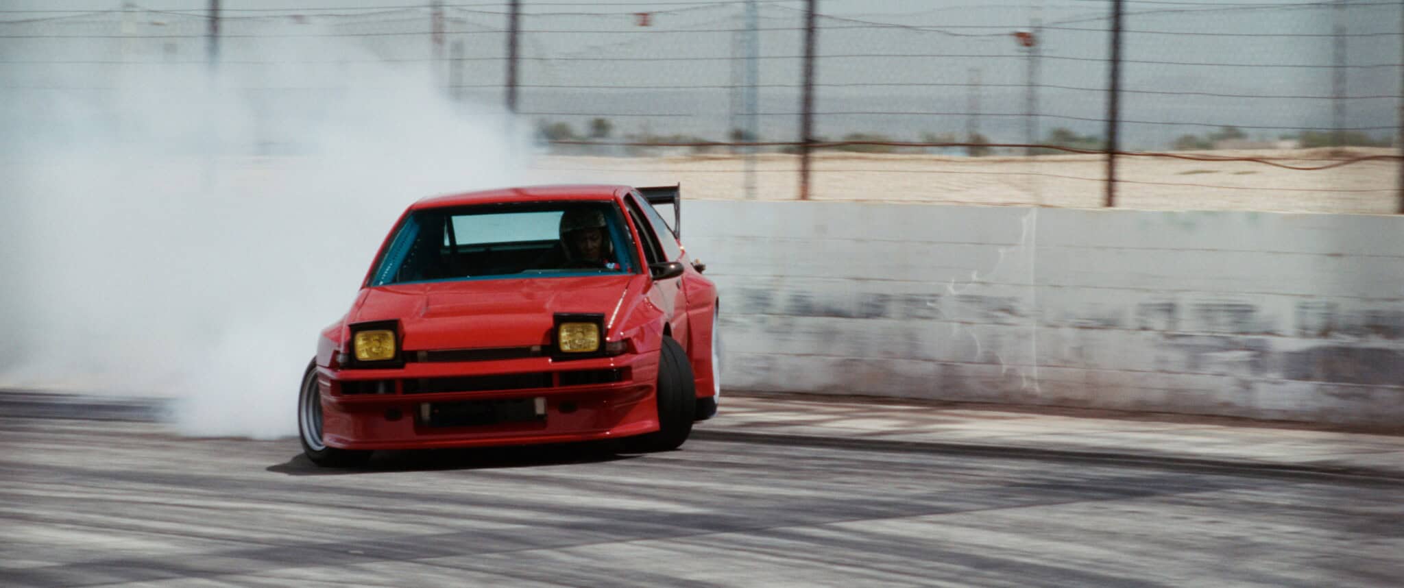 A red sports car drifts around a racetrack corner, creating a large cloud of tire smoke. The driver is visible inside, and blurred fencing lines the track in the background.