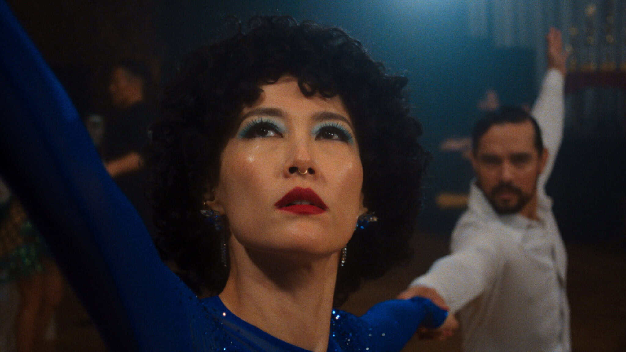 A woman with curly black hair and dramatic blue eye makeup, dressed in a sparkly blue outfit, poses with one arm raised while dancing; a male partner in white is seen blurred in the background.