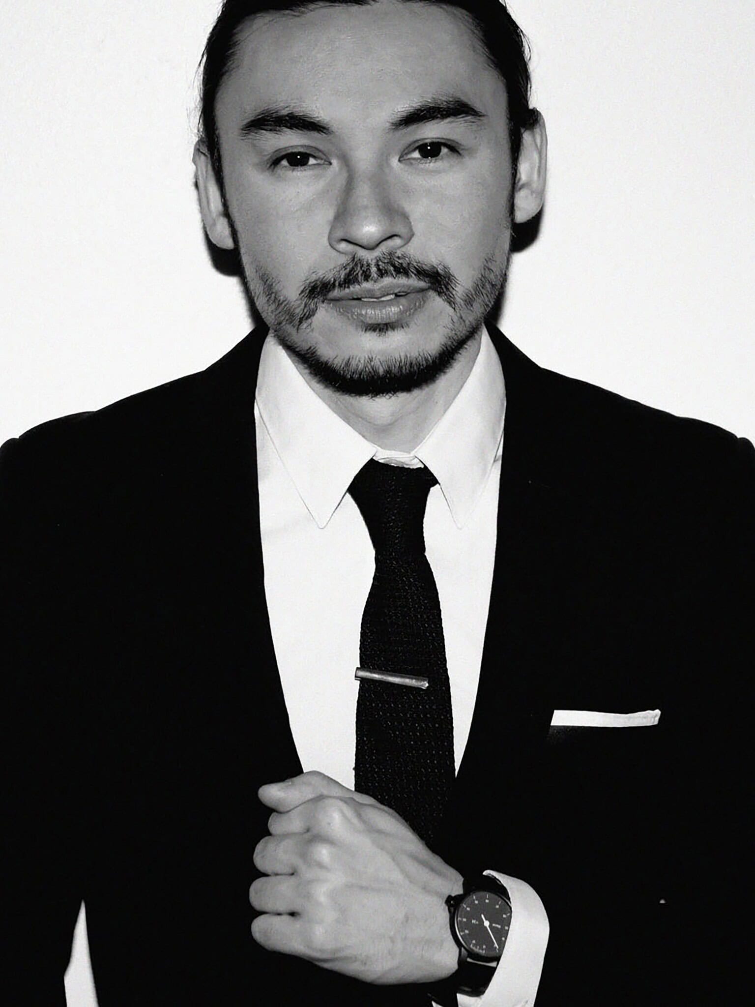 A man in a suit and tie stands against a plain white background, looking at the camera. He has his hand near his chest, wearing a wristwatch, and sports a mustache and beard. The image is in black and white.