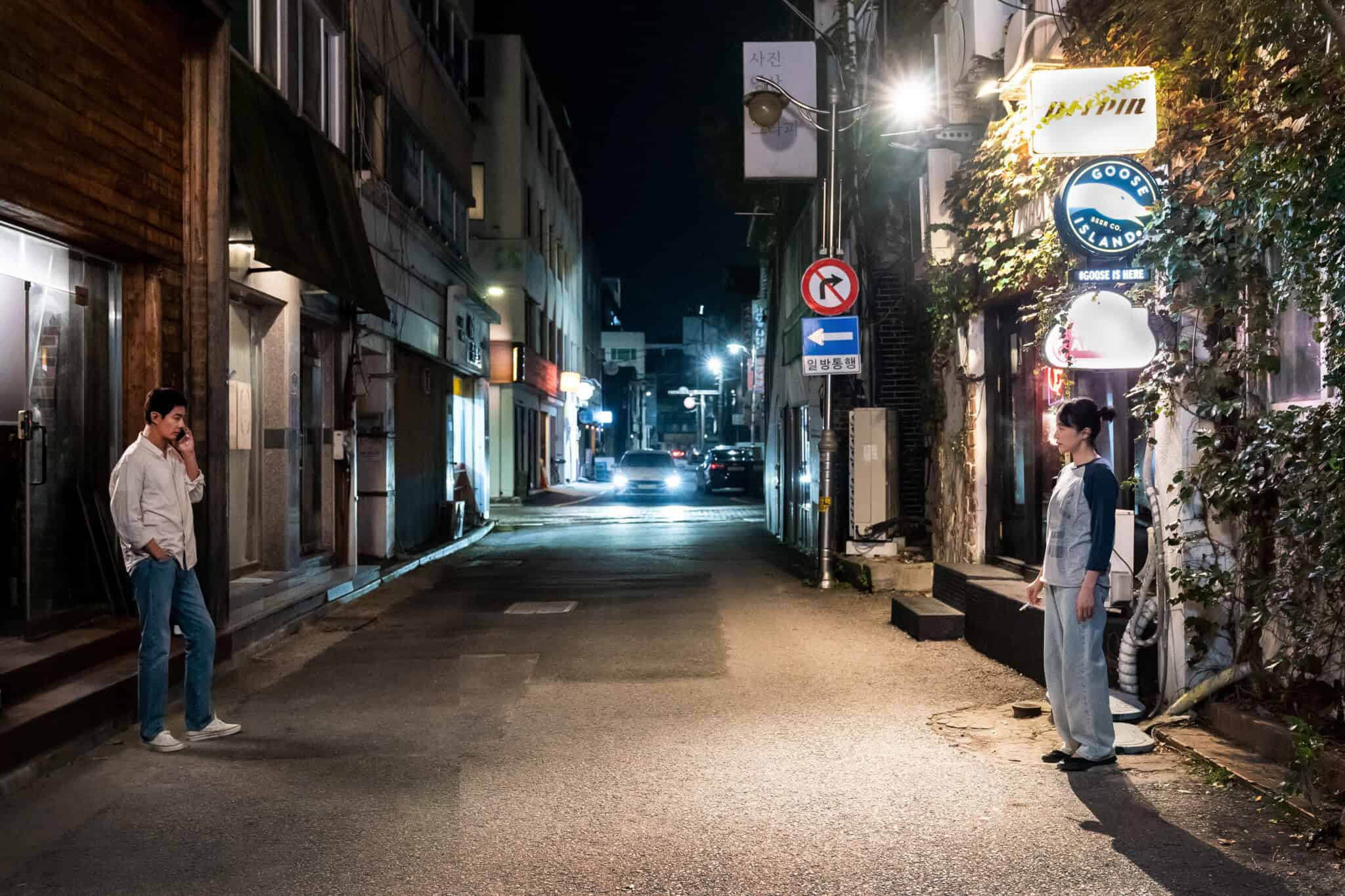 A man and a woman stand apart on opposite sides of a quiet, dimly lit street at night, under glowing shop signs and next to ivy-covered walls.