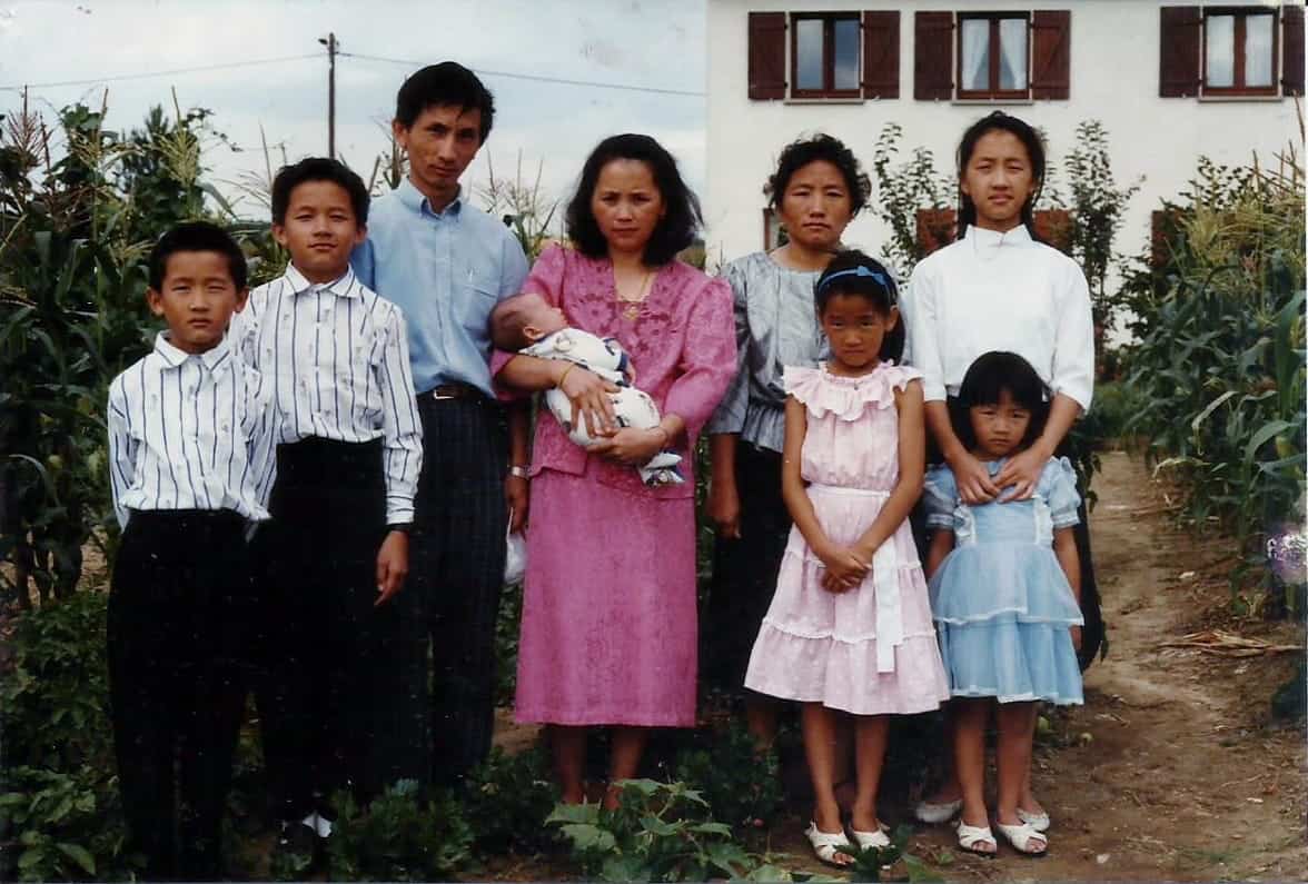 A family of eight, including a baby, stands together outdoors in a garden. The adults and children are dressed formally, and a white building with brown shutters is visible in the background—creating a picture-perfect moment reminiscent of the elegant gatherings often captured by Marc Heu.